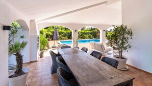 En overdækket terrasse med et træbord og fletstole, en pool i baggrunden og plantekasser med planter.