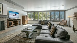 A modern living room with a sectional sofa, a flat-screen TV, a fireplace, and large windows offering a view of trees. - Decatur 1782 (Keystone)
