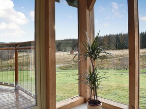 Un balcone in legno con un vaso di fiori, ringhiera in vetro e vista su un prato e alberi.