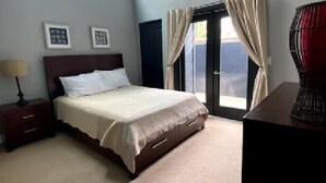 A bedroom with a bed, a nightstand, a lamp, a dresser, and a window with curtains. - Surfrider at the Del Mar Beach Colony (Del Mar)