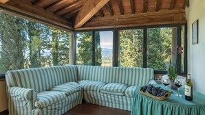 A room with a green-striped sofa, a table with a basket of grapes and wine glasses, and a view of trees and mountains through large windows. - Villalta 18+2, Emma Villas (Arezzo)