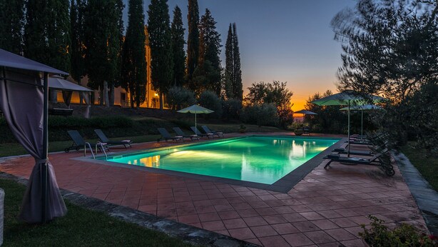 A rectangular swimming pool with a tiled deck, surrounded by lounge chairs and umbrellas, set against a backdrop of trees and a sunset. - Villalta 18+2, Emma Villas (Arezzo)