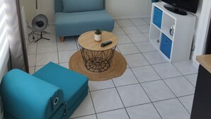 A compact living space with a sofa, a small round table with a plant, a TV on a cabinet, and a fan. - Charming T1 Bis 50m from the Beach and the Market (Pornichet)