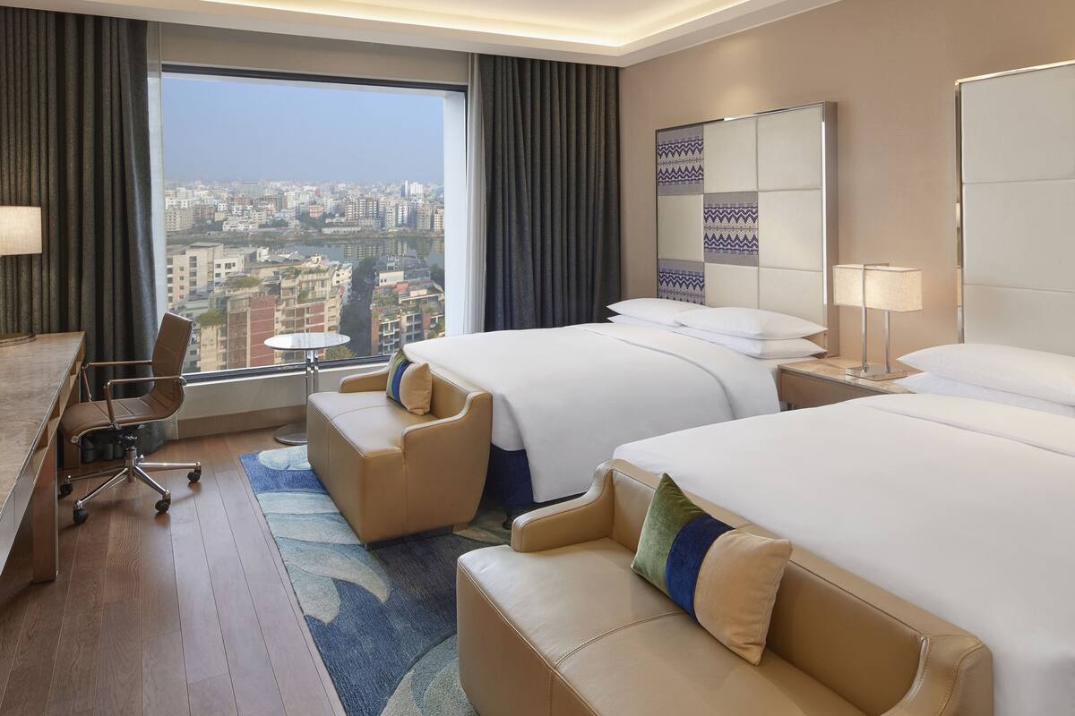 A hotel room with two beds, a desk, a chair, and a view of the city.
