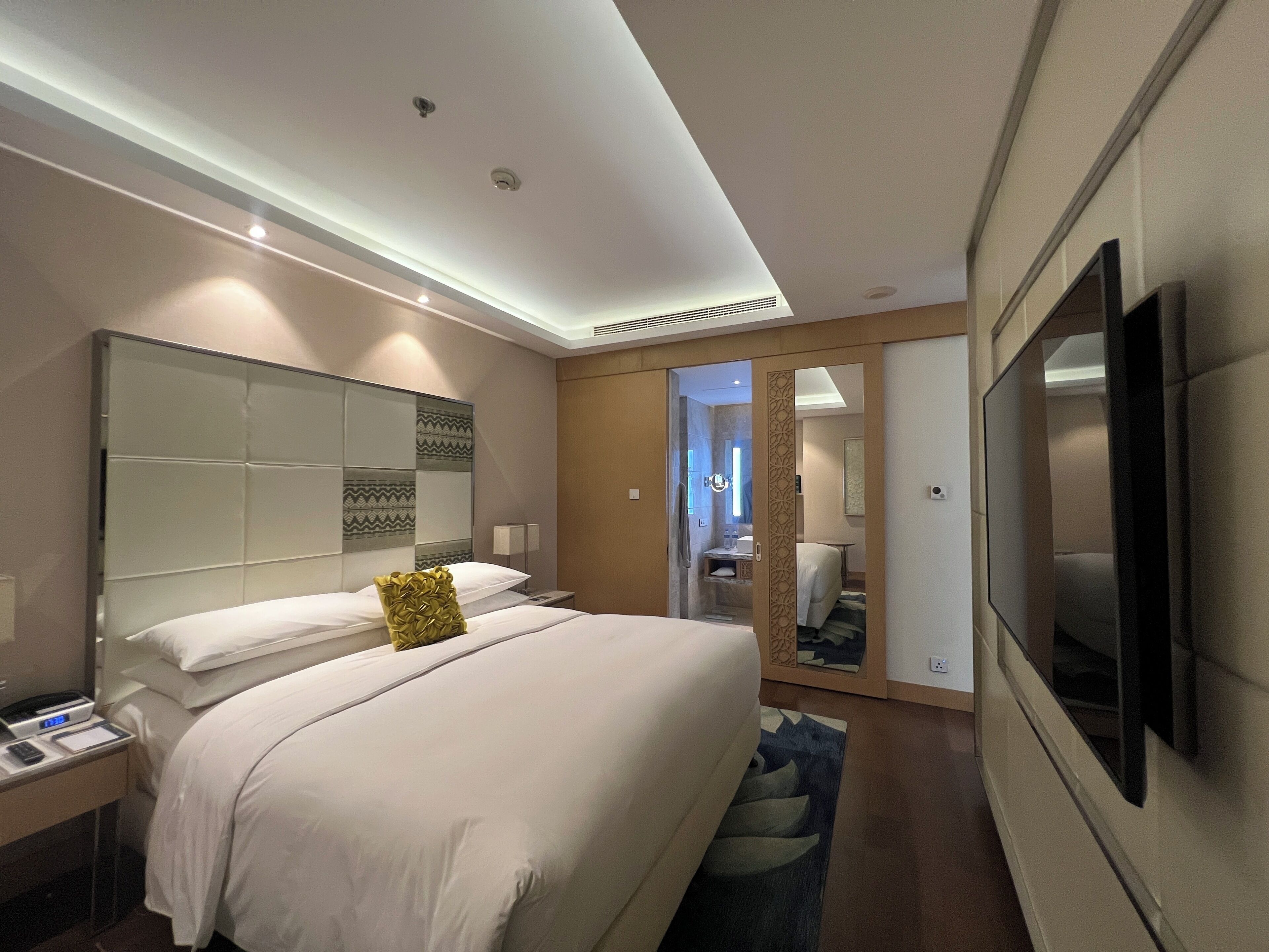 Habitación de hotel moderna con una cama grande, un televisor de pantalla plana, una mesita de noche con lámpara y un baño visible a través de una puerta abierta.
