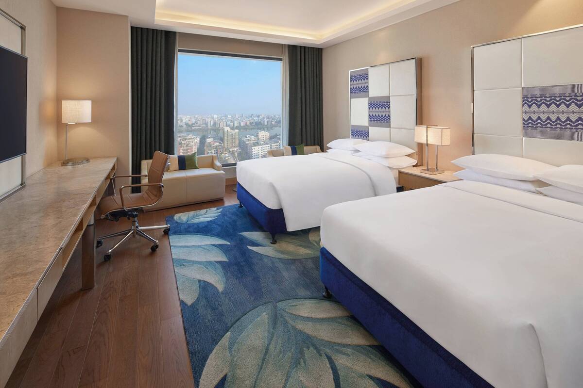 A hotel room with two beds, a desk, a chair, a TV, and a large window with a city view.