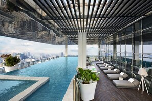 Une piscine à débordement située sur le toit, offrant une vue sur la ville, ainsi qu’un espace de détente avec des sièges et des plantes en pot.