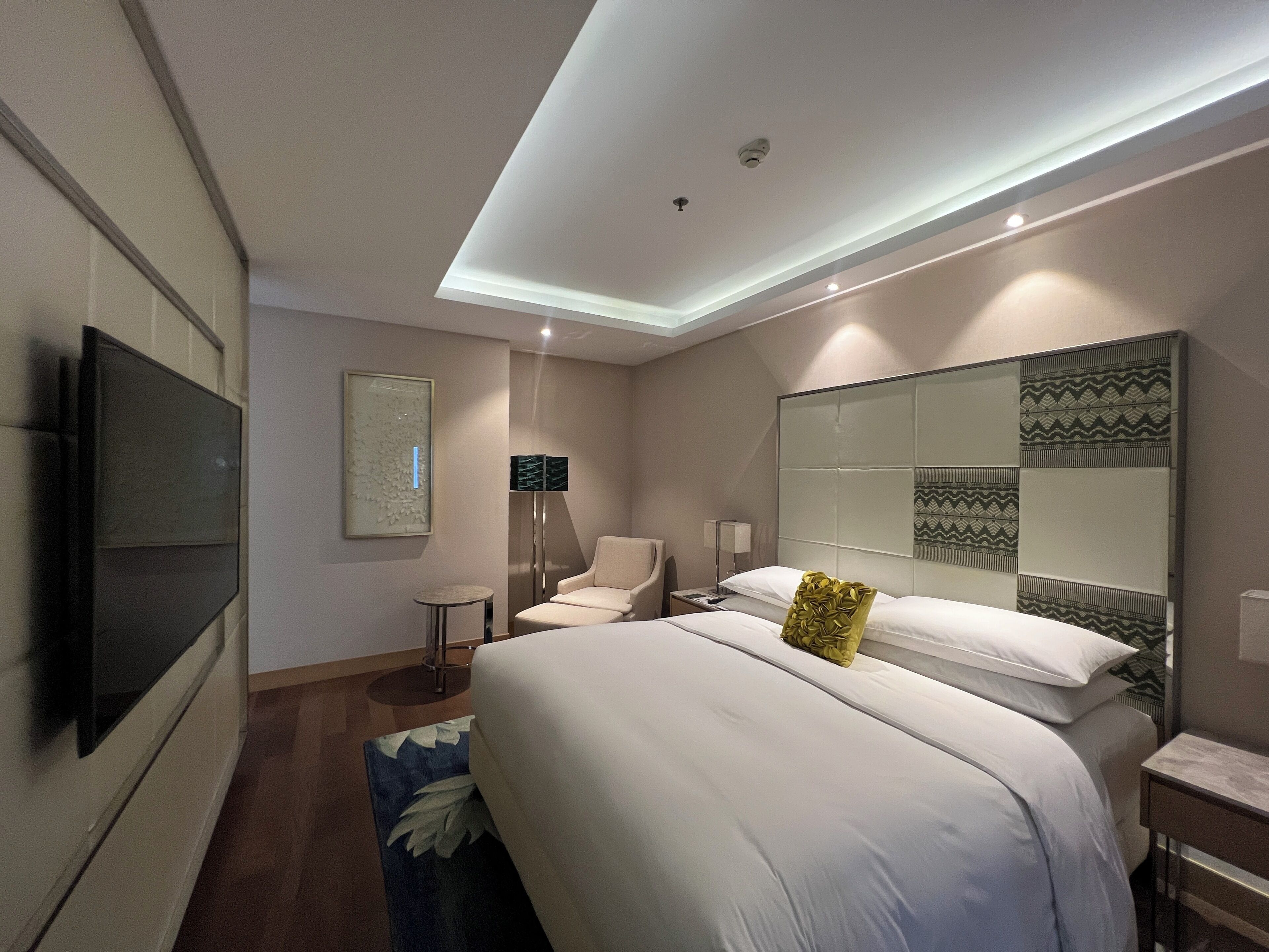 Une chambre d’hôtel moderne dotée d’un grand lit, d’une télévision à écran plat, d’un coin salon avec un fauteuil et d’un miroir décoratif.