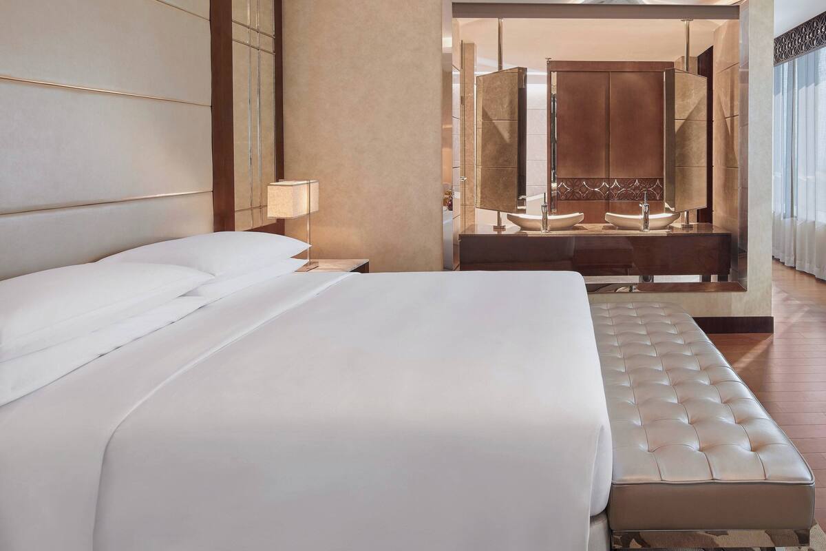 A modern hotel room with a large bed, a bedside table with a lamp, a bathroom with a large mirror, and a view of the city.