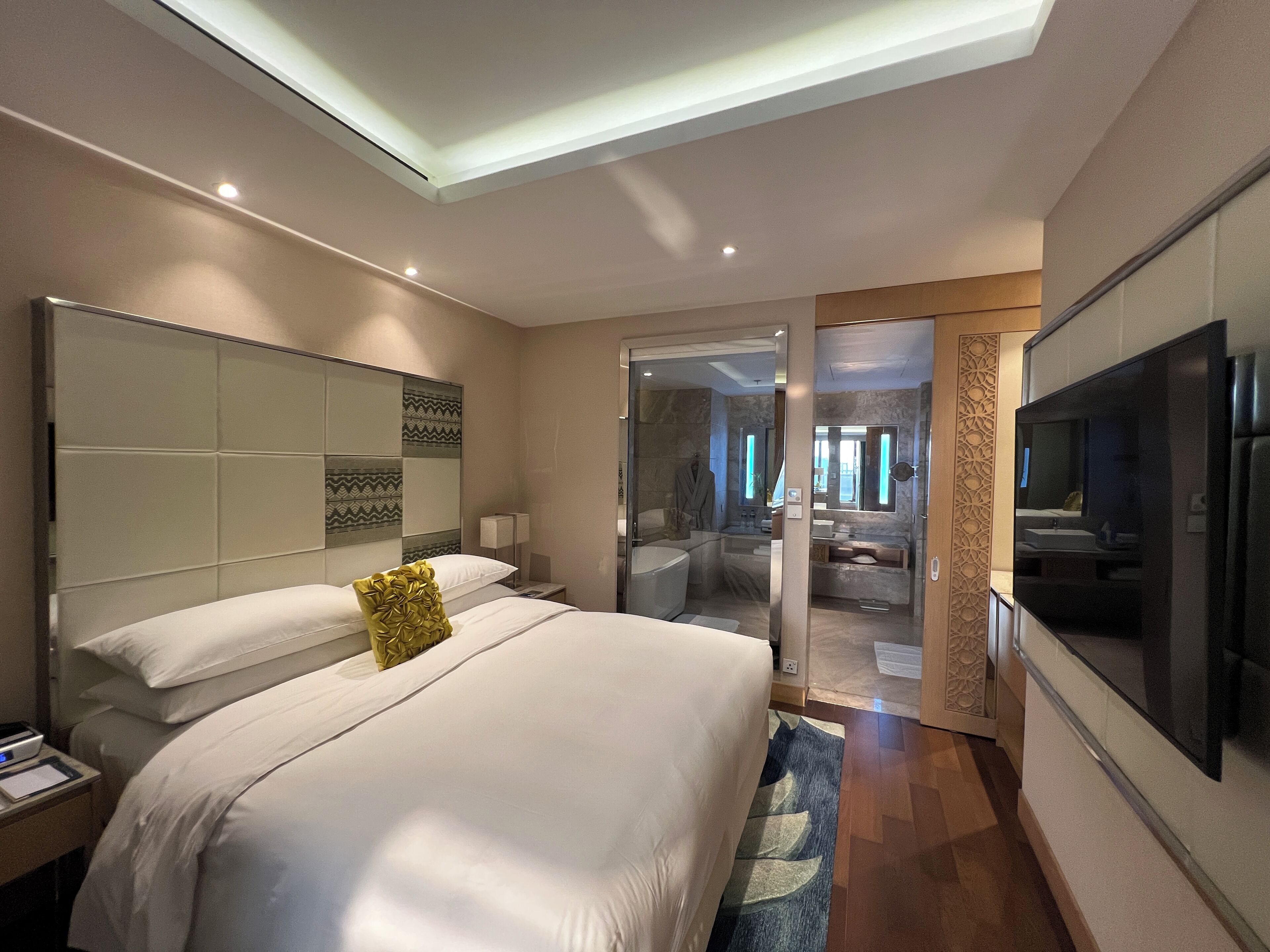 A modern hotel room with a large bed, a flat-screen TV, a bathroom with a bathtub, and a glass door leading to a balcony.