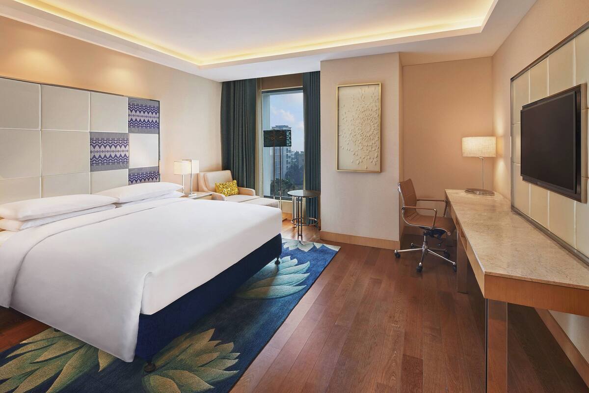 A hotel room with a large bed, a desk, a chair, a TV, and a view of the city through the window.