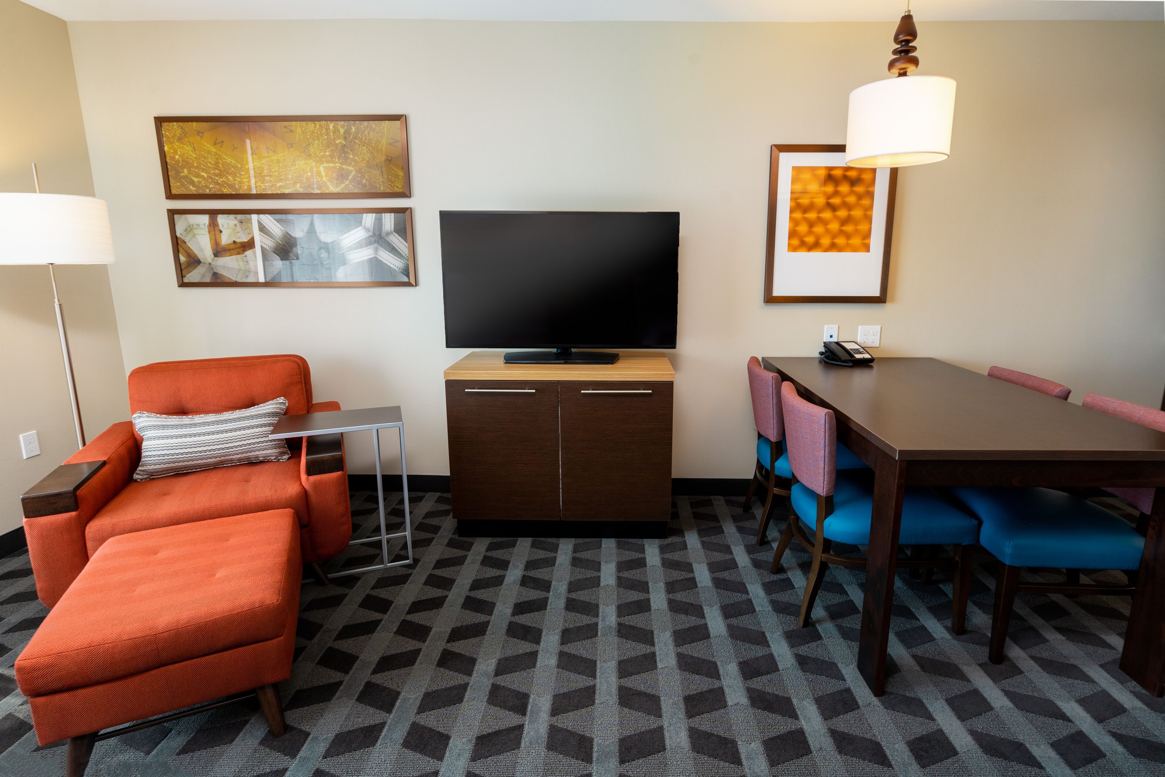 Una habitación de hotel moderna con televisor de pantalla plana, un sillón naranja, mesa de comedor con sillas azules y alfombra estampada.