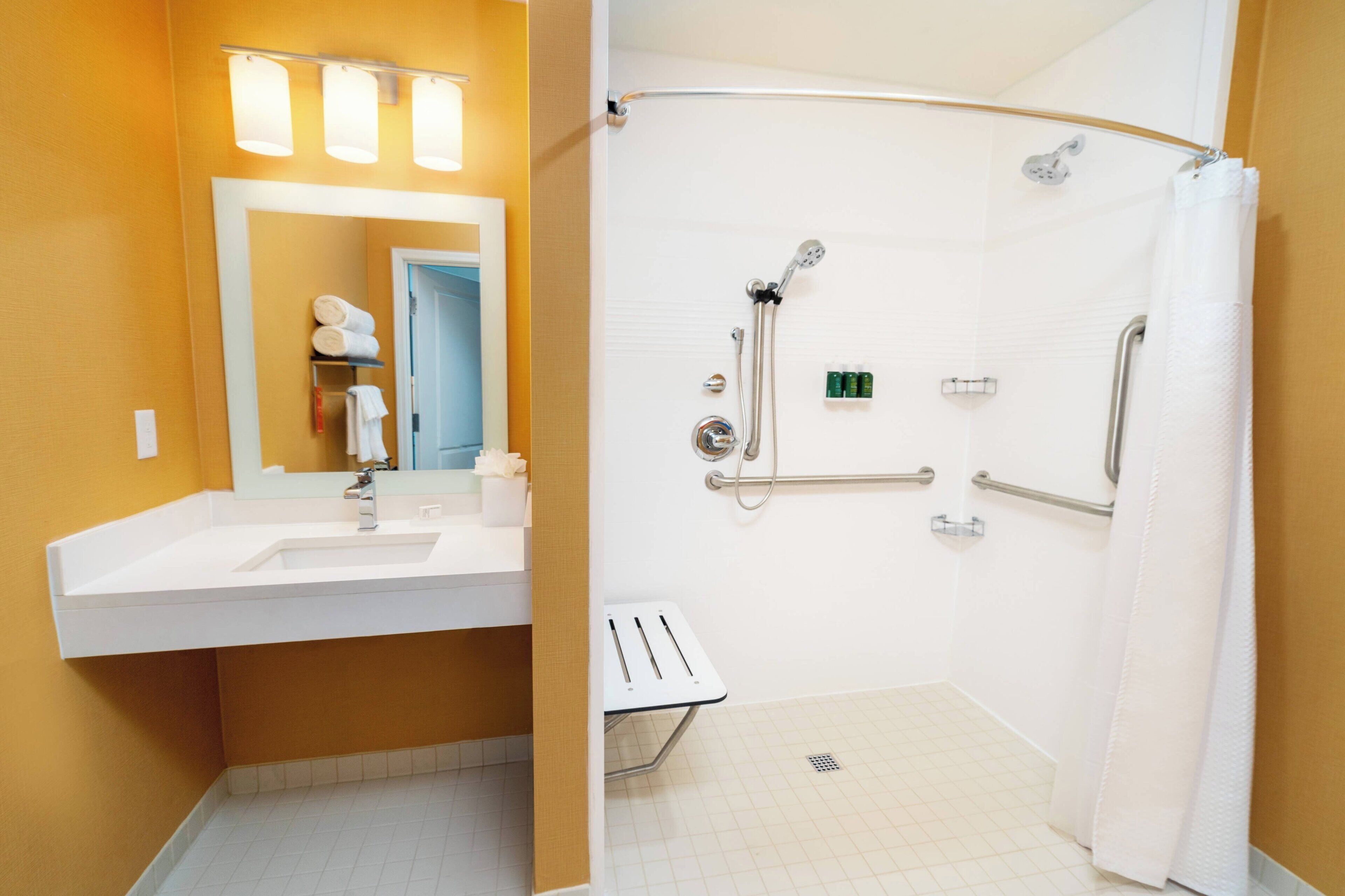 Une salle de bain avec un lavabo blanc, un miroir et une douche équipée de barres d’appui.