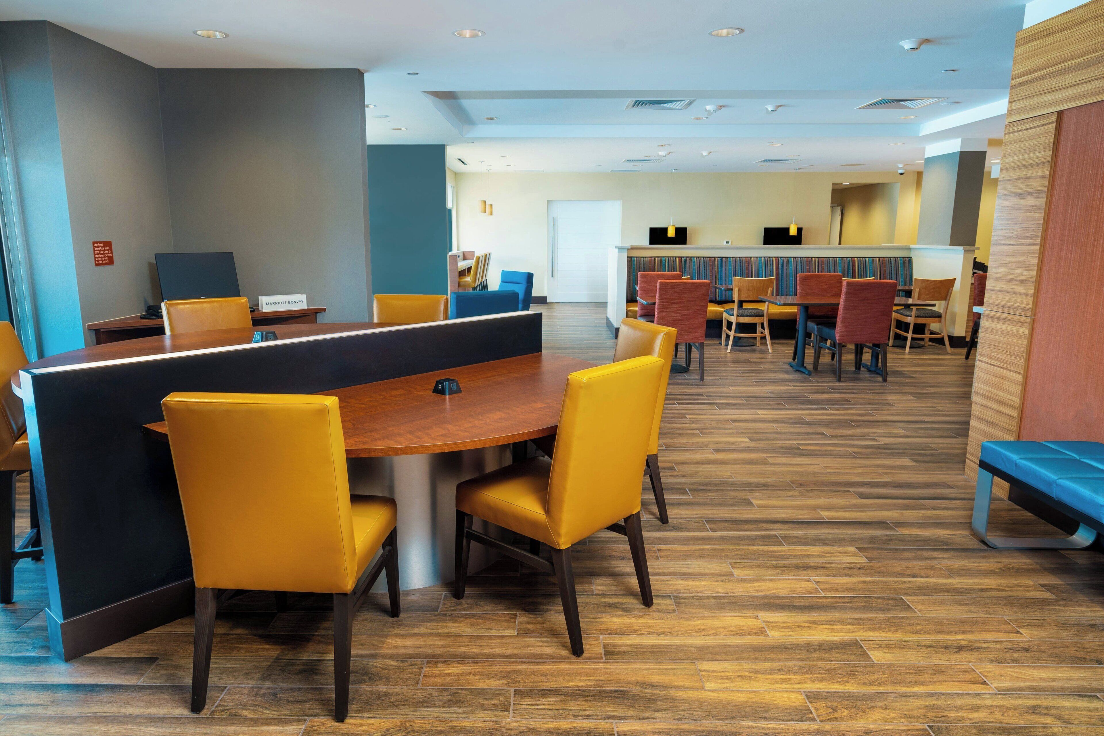 A modern dining area with a wooden table and yellow chairs, a counter with a digital screen, and a wooden floor.