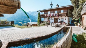 Un edificio alpino tradicional con fachada de madera, un patio con fuente de agua y un telón de fondo montañoso.
