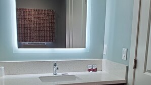 Ein Badezimmer mit einem weißen Waschbecken, einem Spiegel darüber und einer Holzanrichte darunter. Handtücher liegen auf der Anrichte und eine Lotionflasche steht auf der Ablagefläche.