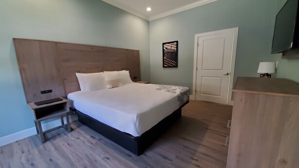 A hotel room with a large bed, a wooden headboard, a bedside table with a phone, a small cabinet, and a door with a light on it.
