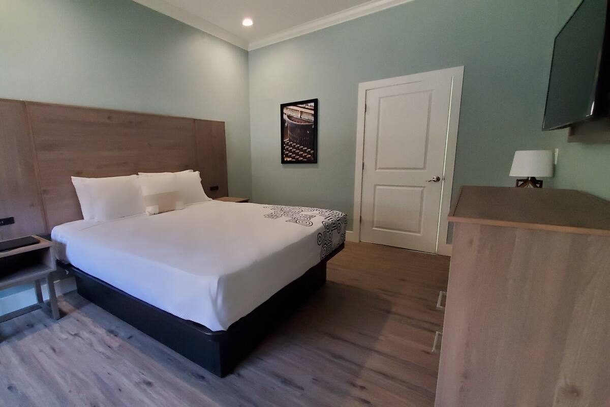 A hotel room with a large bed, a wooden headboard, a bedside table with a phone, a small cabinet, and a door with a light on it.