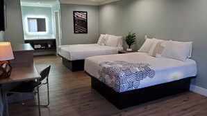 Ein Hotelzimmer mit zwei Betten, einem Schreibtisch mit Stuhl, einer Topfpflanze und einem gerahmten Bild an der Wand.