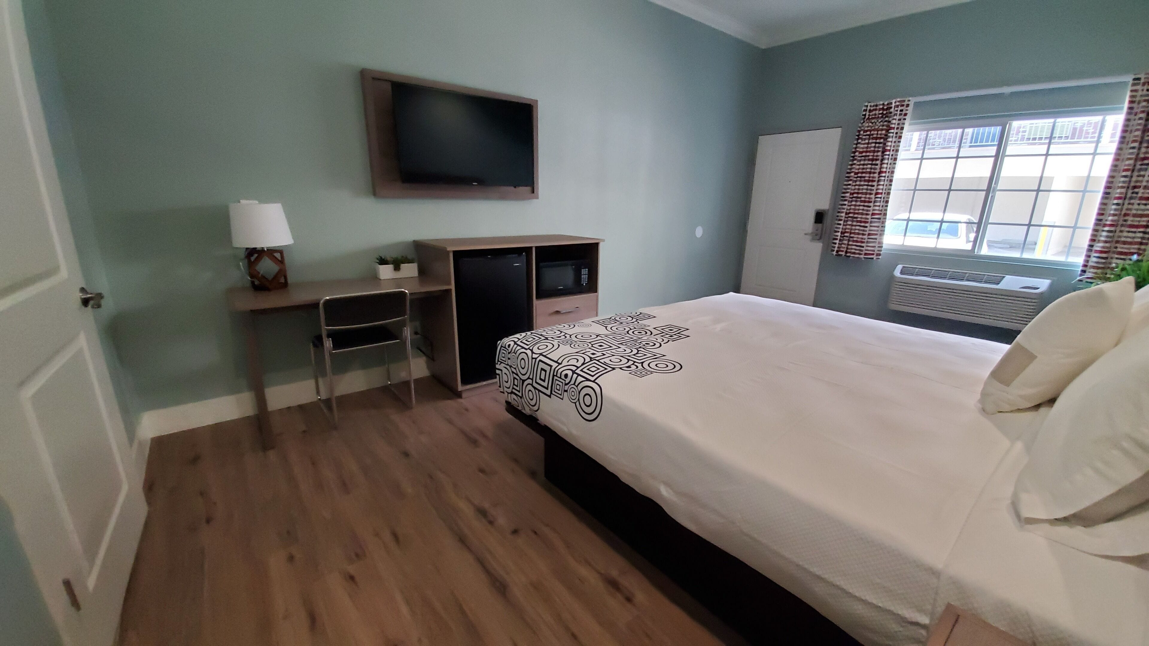 A bedroom with a bed, a desk, a TV, and a window with curtains.
