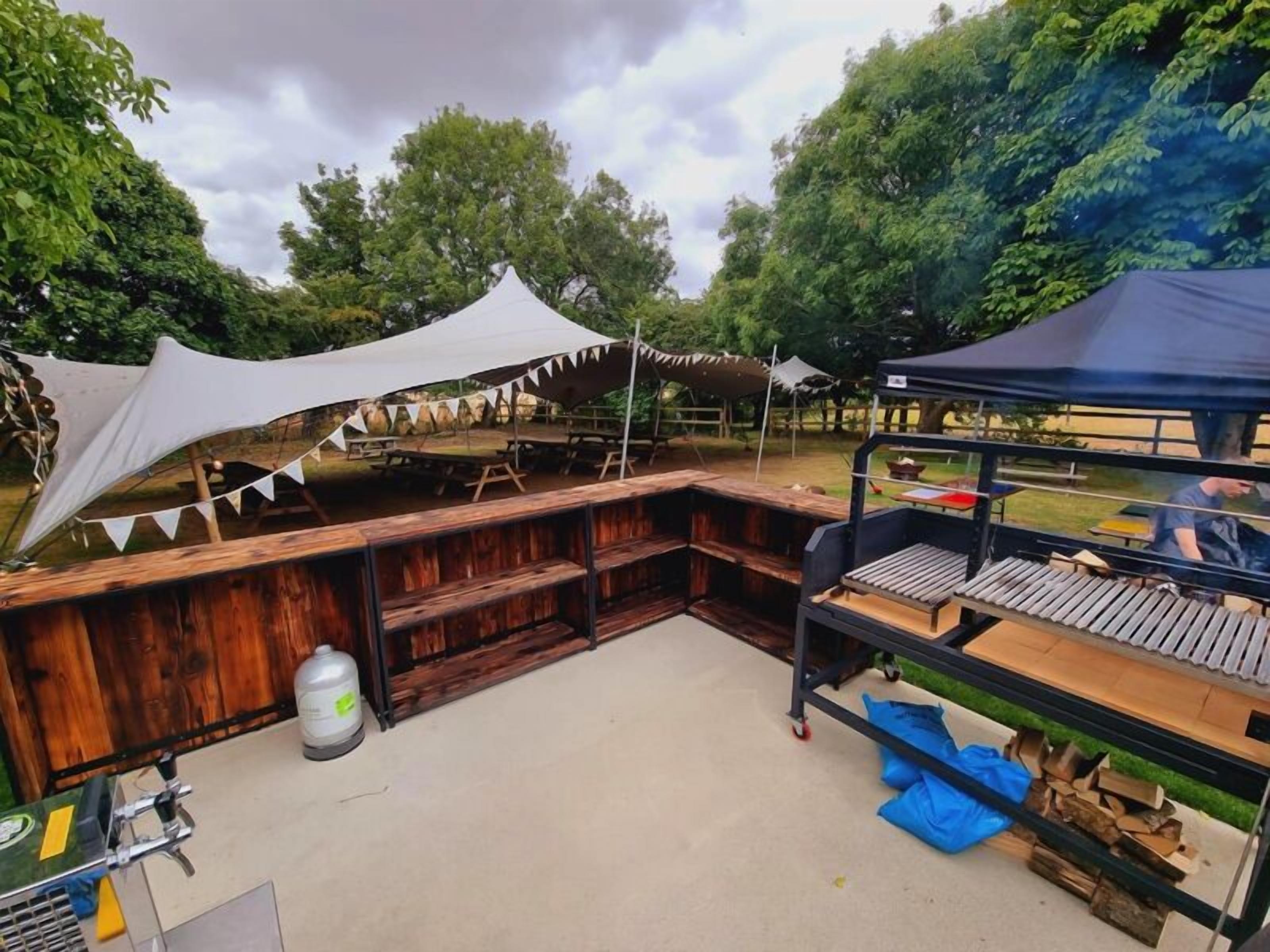 An outdoor barbecue area with a wooden bench and grill, surrounded by trees and covered with a white canopy.