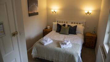 A neatly made bed with white linens and a gray throw blanket, two bedside tables with lamps, a nightstand with a lamp, a wall-mounted picture, and a radiator.