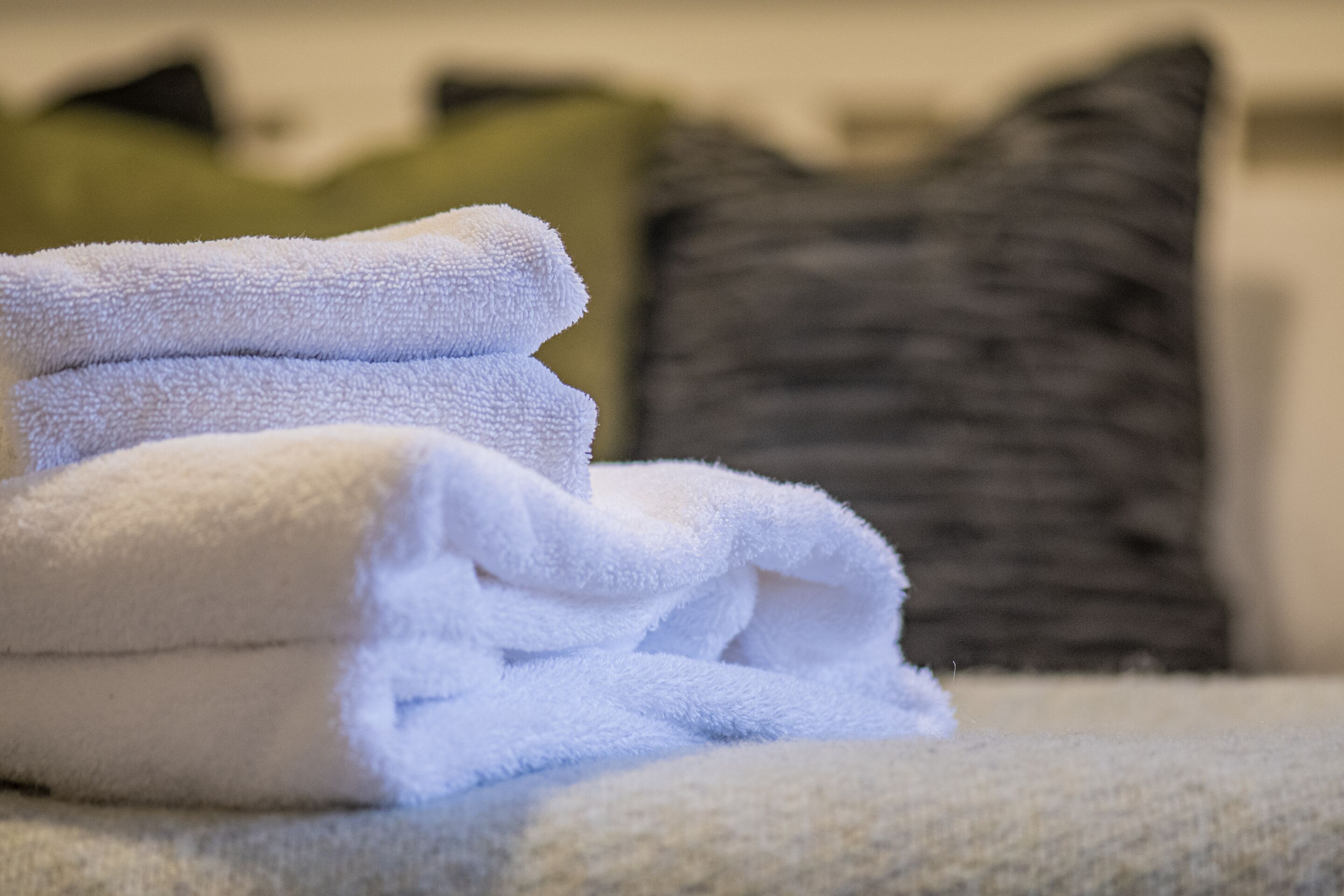 Una pila de toallas blancas sobre una cama, con una almohada gris y verde al fondo.