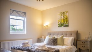 Une chambre à coucher avec un grand lit, une fenêtre ornée d’une lambrequin à motifs de feuilles, un luminaire fixé au mur, une table de chevet et un tableau représentant une scène de forêt.