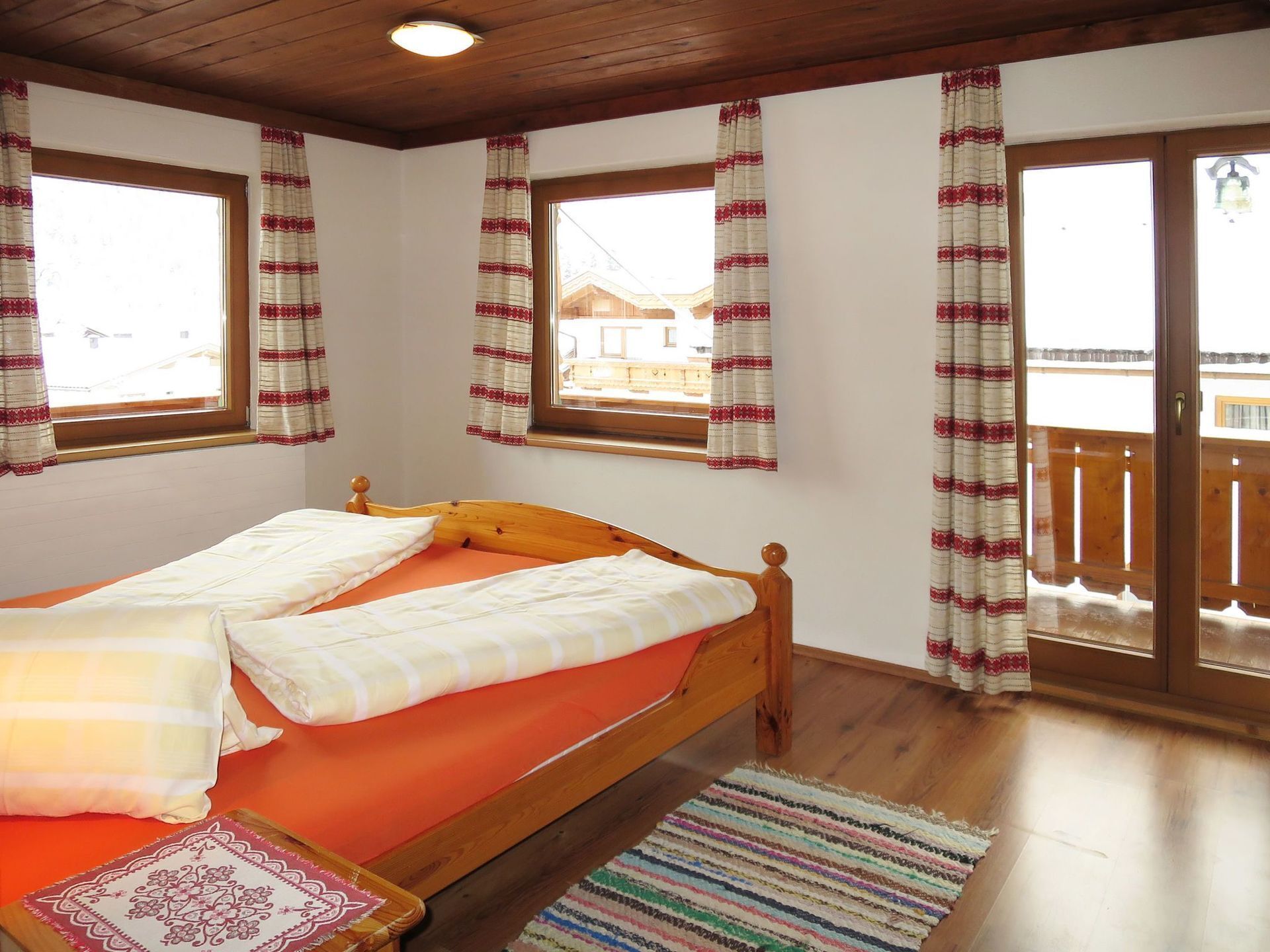 A room with a wooden bed, two windows with curtains, a wooden floor, and a balcony with a railing.