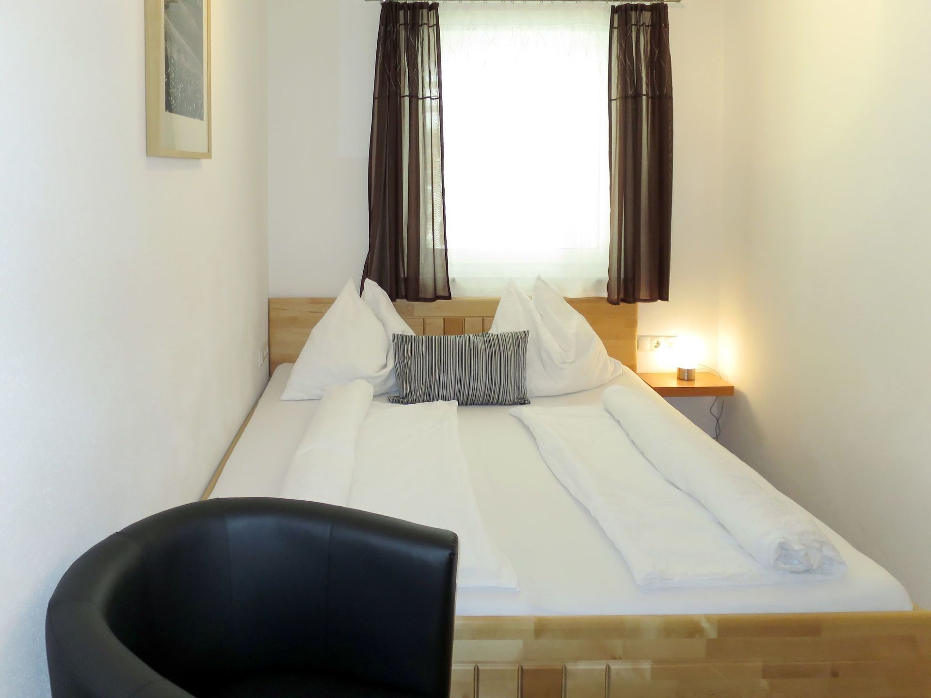 A neatly made bed with pillows and a striped pillow, a bedside table with a lamp, and a black chair in a simple room with a window and curtains.