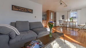 A modern living room with a grey sofa, a coffee table with a magazine, a vase with flowers, and a dining area with a wooden table and chairs.