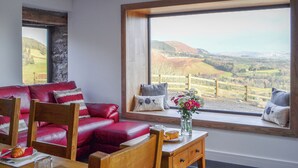 Une pièce chaleureuse avec un canapé en cuir rouge, une table en bois sur laquelle se trouve une assiette de nourriture et un verre, et une grande fenêtre offrant une vue panoramique sur des collines verdoyantes.