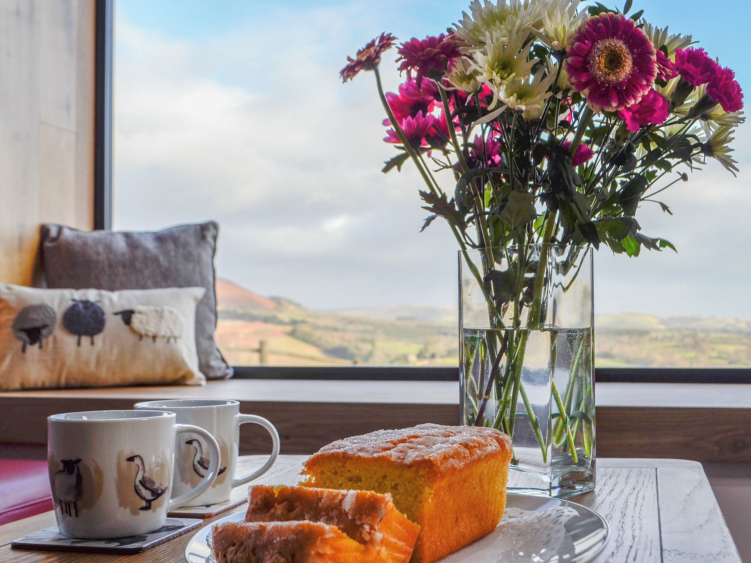 A slice of cake on a plate, a mug with a sheep design, and a vase with flowers by a window.