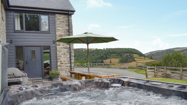 Un espace avec un bain à remous, une table et un banc en bois, un mur en pierre et une vue sur une maison et des collines en arrière-plan.