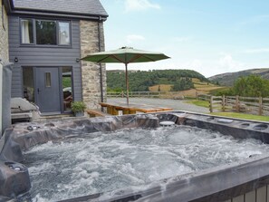 A hot tub area with a wooden table and bench, a stone wall, and a view of a house and hills in the background.