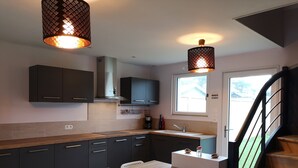 A modern kitchen with dark cabinets, a wooden countertop, a sink, and a dining table with chairs. There are pendant lights above the counter and a window with a view outside.