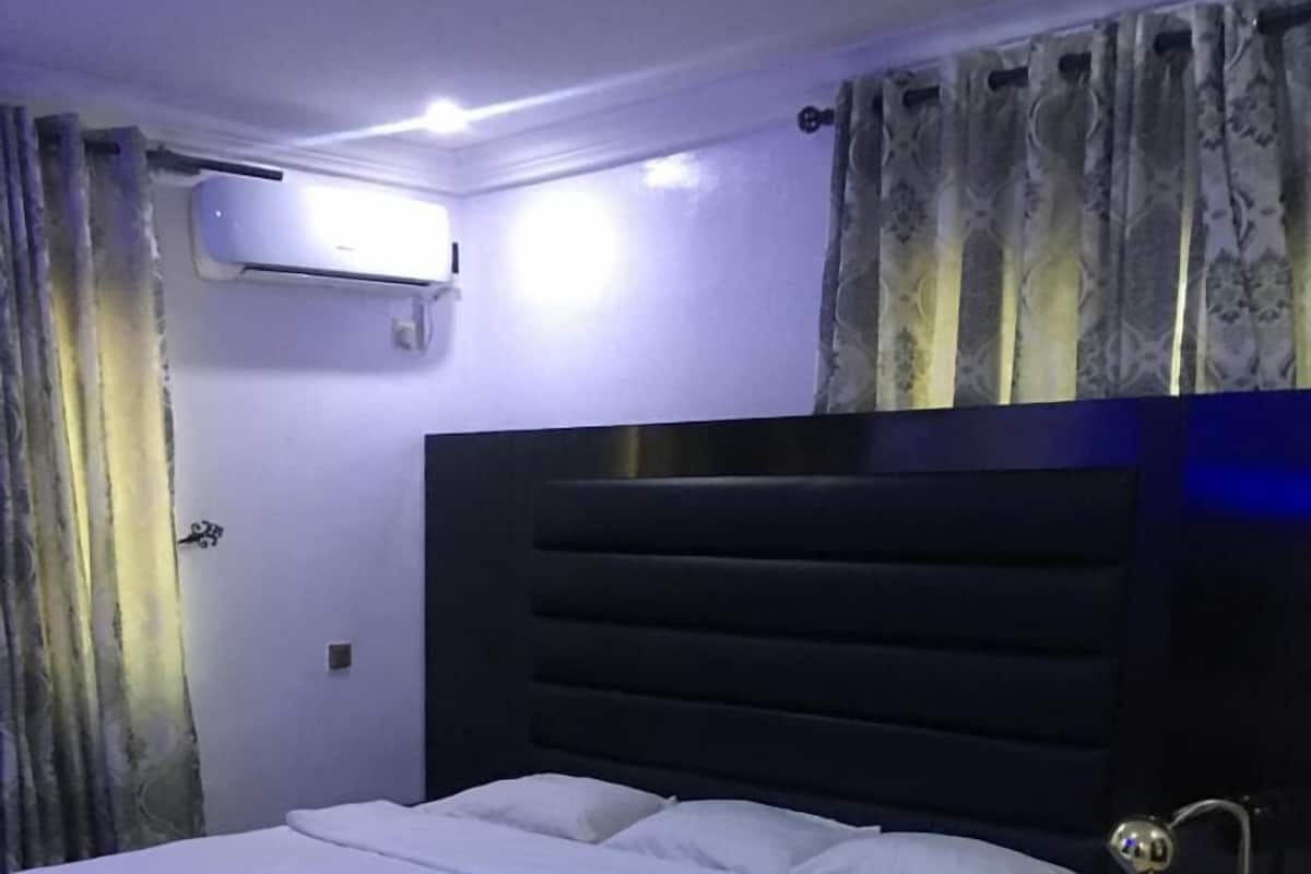 A bedroom with a large bed, a nightstand, a lamp, and a wall-mounted air conditioner.
