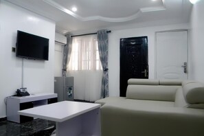 A modern hotel room with a flat-screen TV mounted on the wall, a white desk, a beige sofa, and a door with a decorative handle. - Jaybritt Luxury Apartment (Ibadan)