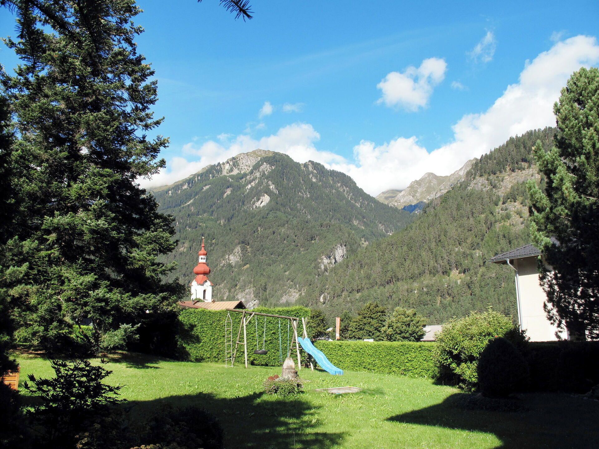 Ein Rasen mit Schaukeln, ein kleines Gebäude mit rotem Dach und einem Berg im Hintergrund.