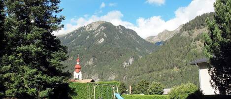 Ein Rasen mit Schaukeln, ein kleines Gebäude mit rotem Dach und einem Berg im Hintergrund.