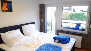A bedroom with a bed, white pillows, a blue quilt, and a window with a view of greenery and a building.