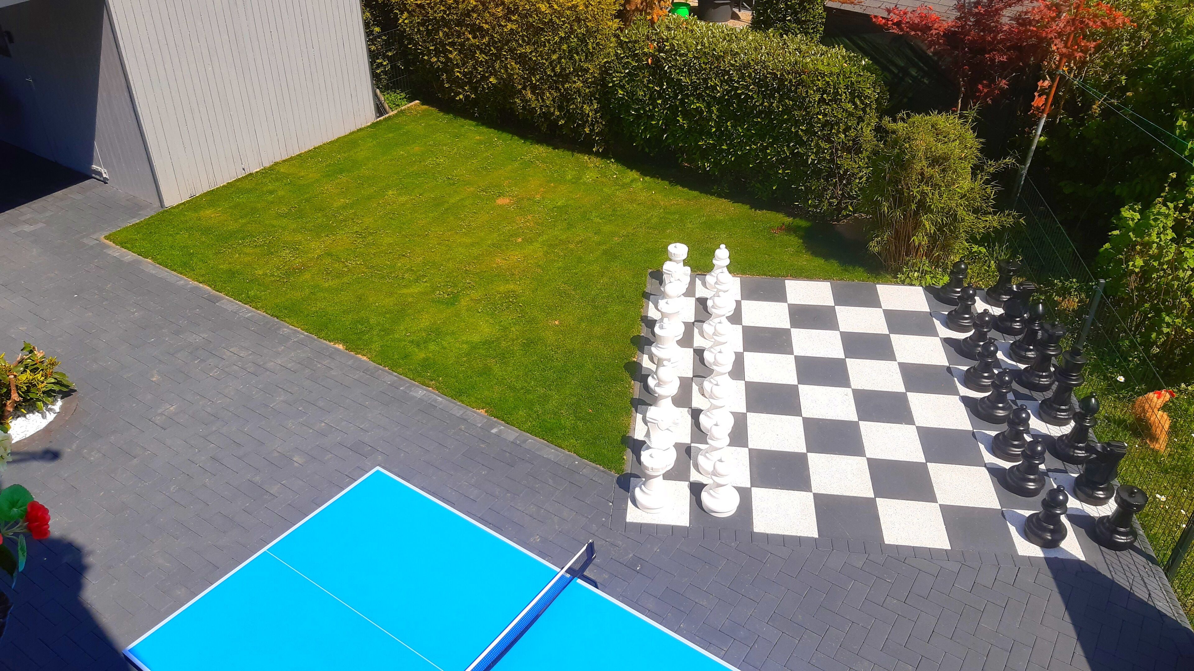 A checkered outdoor chessboard with pieces, a blue pool, and a paved area with a hedge and trees.