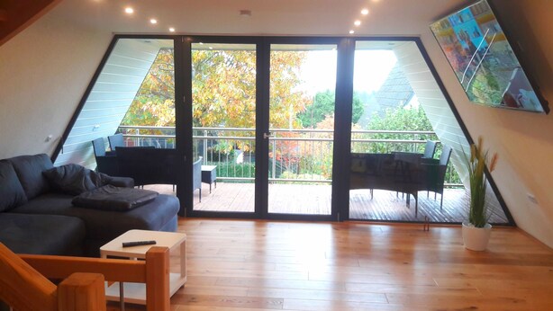 A spacious loft bedroom with a large sliding glass door leading to a balcony, a dark-colored sofa, a small wooden table, and a potted plant.
