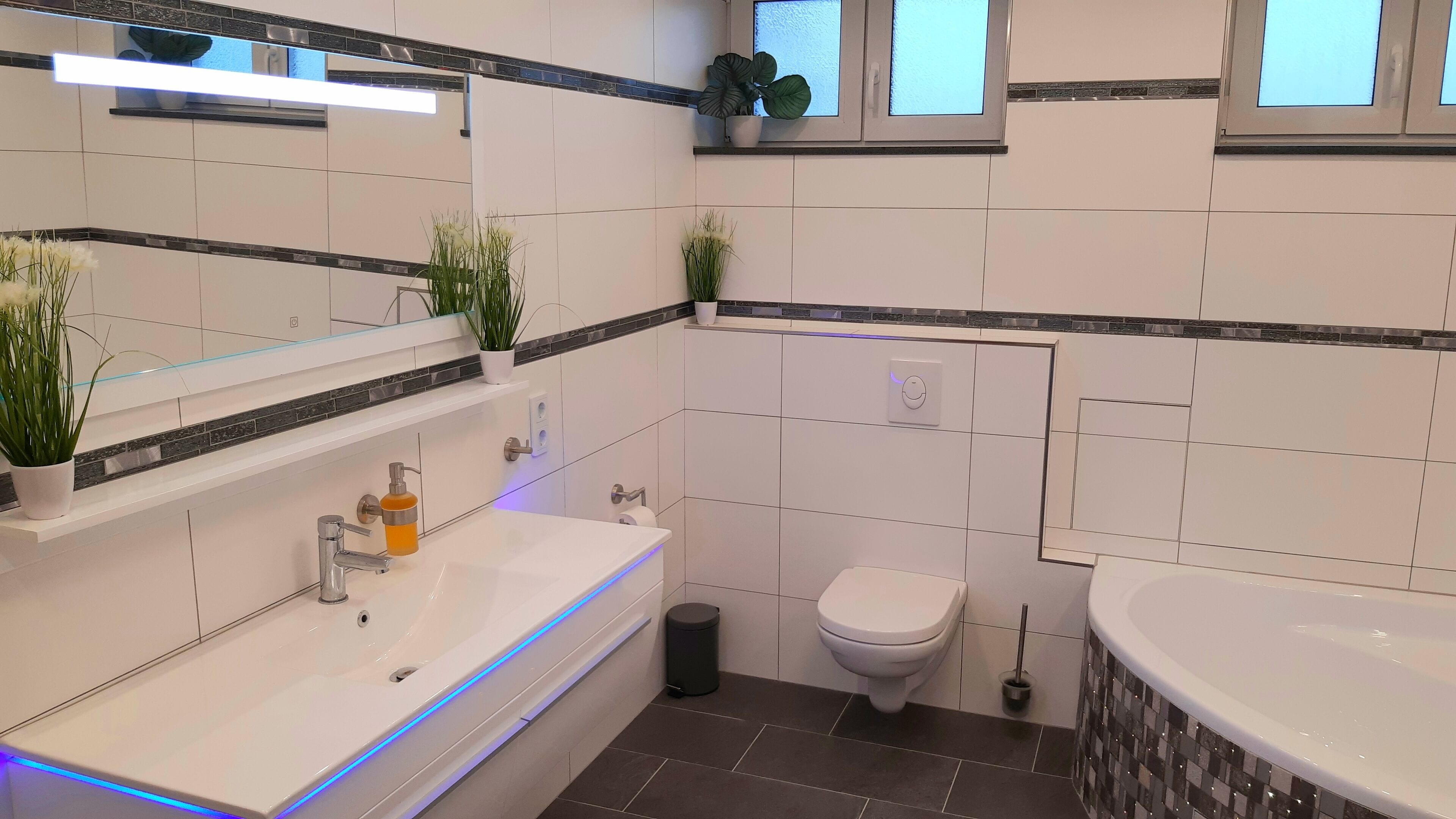 Une salle de bain moderne avec un lavabo blanc, des toilettes et une baignoire. Des plantes en pot sont posées sur le comptoir et une fenêtre se trouve au-dessus des toilettes.