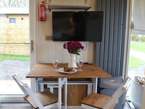 Un petit coin repas avec une table en bois, deux chaises, un vase blanc orné de fleurs et un téléviseur fixé au mur.