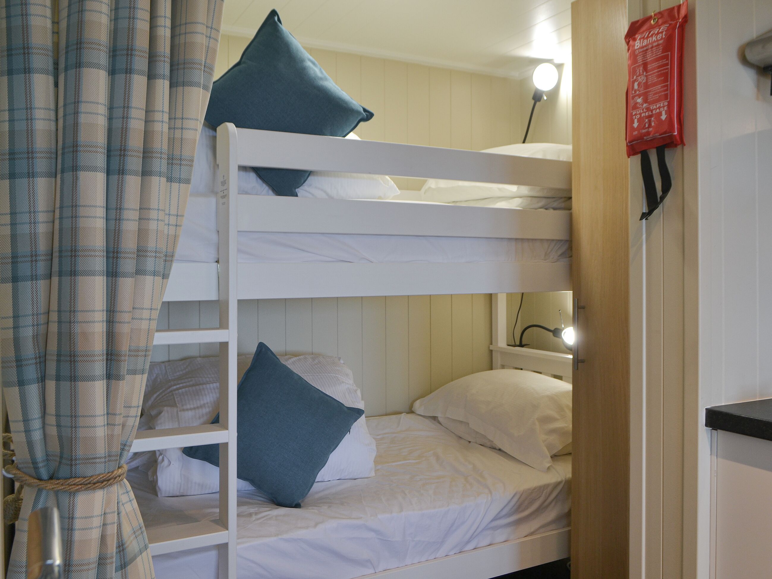 Un lit superposé avec une literie blanche et des oreillers bleus, une lampe de chevet et une couverture anti-feu accrochée au mur.