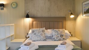 Een netjes opgemaakt bed met bloemenkussens, een bedlampje en een klok aan de muur.