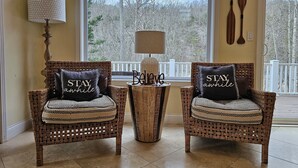 Two wicker chairs with patterned cushions, a small round table with a lamp, and a view of trees through a large window. - Welcome to Hidden Haven – A Spacious Lakeside Retreat (Burnside)