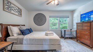 Un dormitorio con cama, ventilador de techo, televisión, cómoda y ventana con vistas a la vegetación.