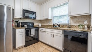Una cocina moderna con armarios blancos, electrodomésticos de acero inoxidable, encimeras de granito y un ventanal con persianas.